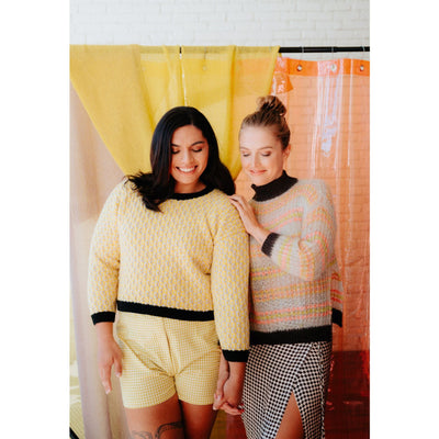 Two women stand together holding hands and smiling while wearing their knit sweaters.