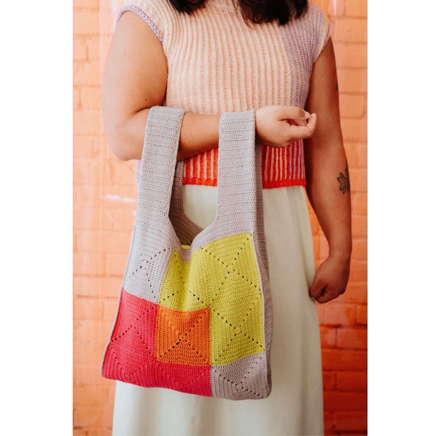 A woman in a short sleeve knit sweater stands holding a colorful crochet handbag. 