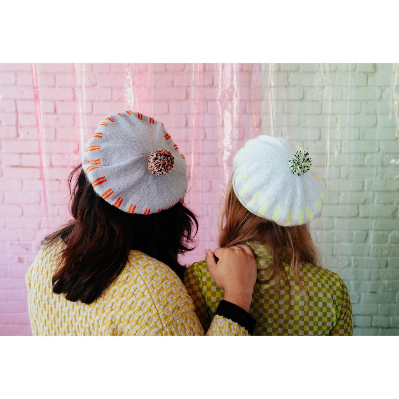 Two women stand together wearing knit beret hats in neutral shades and featuring bright pops of color. 