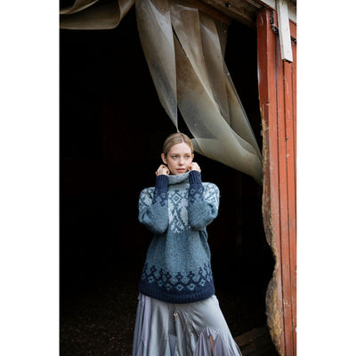 Model standing in rustic doorway wearing Iku Turso sweater knit with blue shades of Rauma Vams. This is a design from the Knitted Kalevala book by Jenna Kostet and published by Laine.