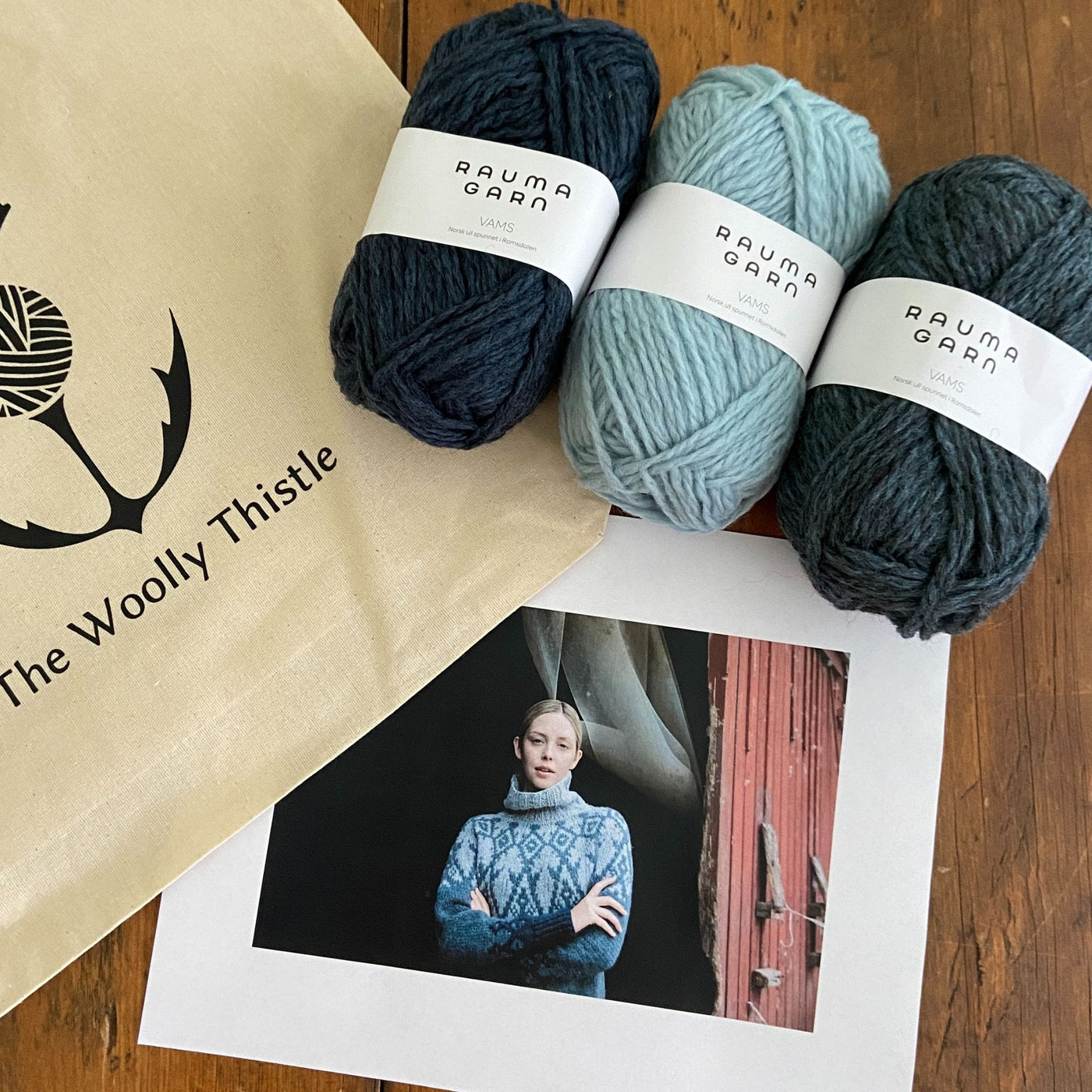 Components of the Iku Turso sweater yarn set knit with blue shades of Rauma Vams. This is a design from the Knitted Kalevala book by Jenna Kostet and published by Laine. Three shades of Rauma Vams yarn on table with photo of sweater and The Woolly Thistle Tote Bag. 