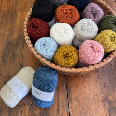 A basket of skeins of Rauma Gammelserie on a wooden table, with skeins of natural 401 and blue 438 nearby