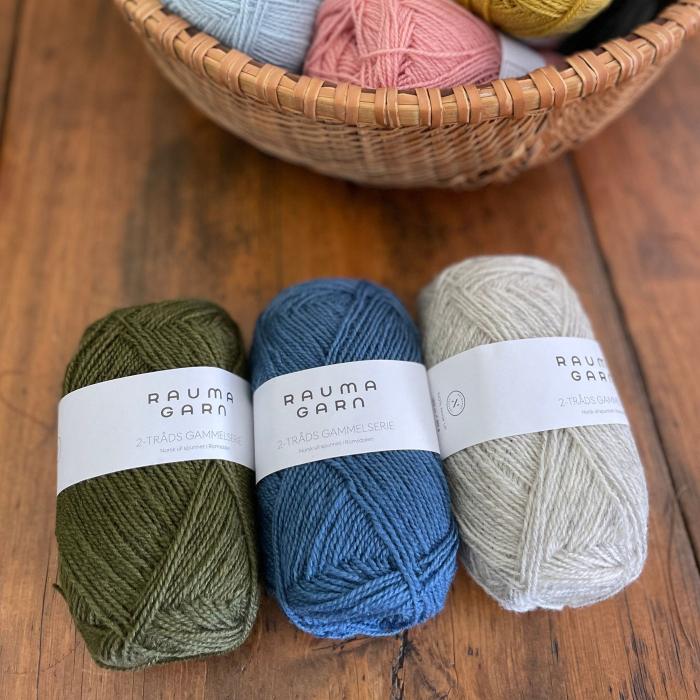 Three skeins of Rauma Gammelserie in green 476, blue 438, and light grey 403, on a wooden table near a basket