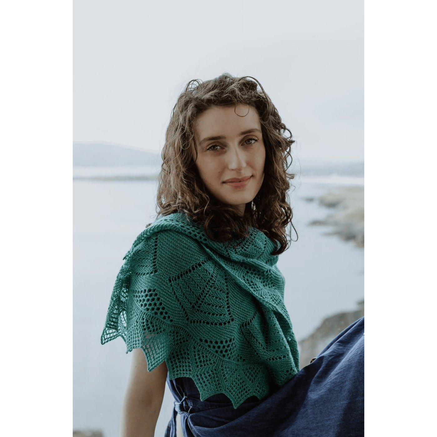 The Woolly Thistle image from Shetland Wool Week Annual 2021 featuring a woman sitting by the ocean wearing knitted green shawl