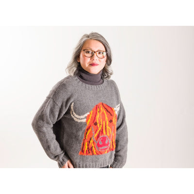 Woman wearing a grey sweater with Highland Cow design knit in orange on front of sweater. 