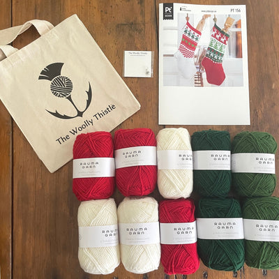 Christmas Stocking Yarn Kit components to make 2 different variations of Stockings. arranged on wooden table. 10 balls of Strikkegarn yarn in red, greens, and white with pattern card and The Woolly Thistle tote and stitch marker.