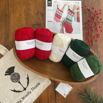 Christmas Stocking Yarn Kit components arranged on wooden table. Strikkegarn yarn in red, green, and white with pattern card and The Woolly Thistle tote and stitch marker. 
