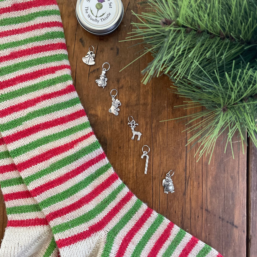 TWT Holiday stitch marker set. Charms lined up vertically along a pair of Stockings knit with WYS Candy Cane yarn. Charms include bells, snoman, stocking, reindeer, candy cane, and santa. TWT tin is shown at top. 