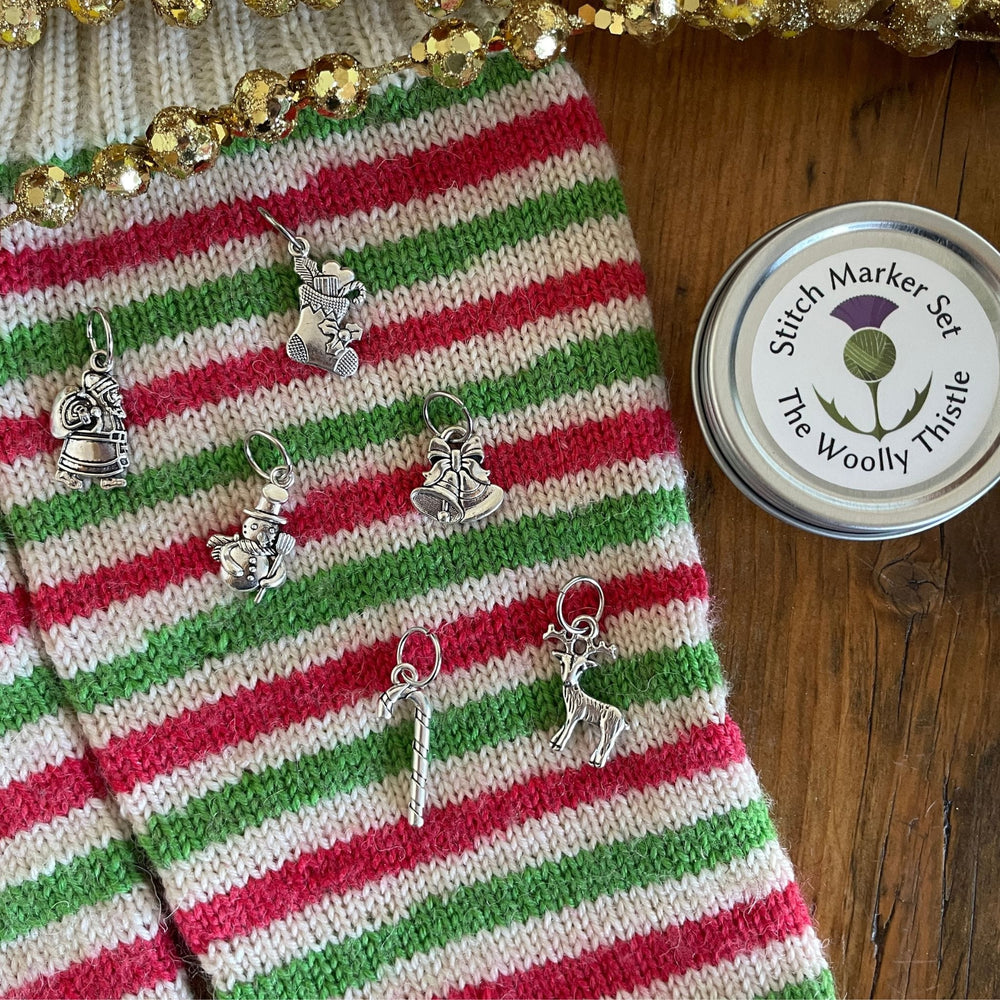 TWT Holiday stitch marker set. Charms arranged atop a pair of Stockings knit with WYS Candy Cane yarn. Charms include bells, snowman, stocking, reindeer, candy cane, and santa. TWT tin is shown on right.