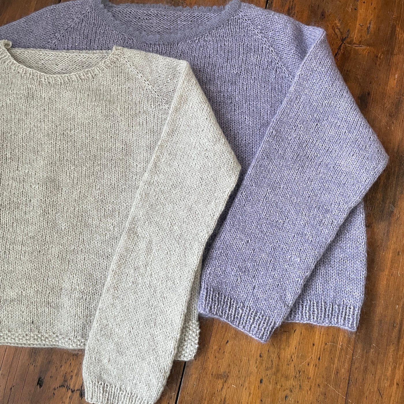 Two sweaters lying on wooden table. Sweaters are The Woolly Thistle Vanilla Fluff pattern in lavender and light grey. 