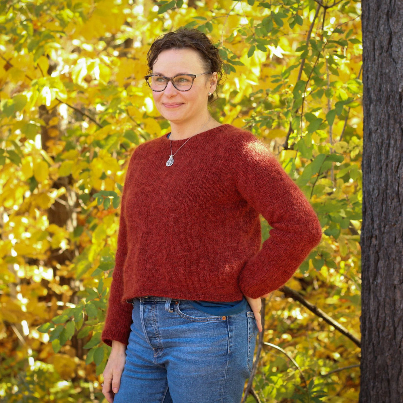 Model standing outside in front of yellow foliage. She is wearing jeans and The Woolly Thistle Vanilla Fluff sweater in color Rauma Finullgarn color 4120 and Rauma Plum 174.