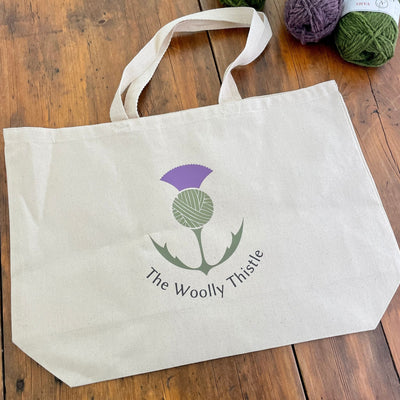 Canvas tote with The Woolly Thistle color logo.  Large size tote with green and purple yarn in background.