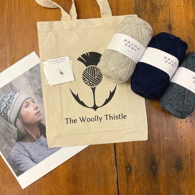 Niobe Hat Yarn Set components shown on wooden background. Three balls of Rauma Finullgarn yarn in two shades of blue and light grey, The Woolly Thistle tote bag and stitch marker alongside a Photo of a woman wearing the knitted hat.