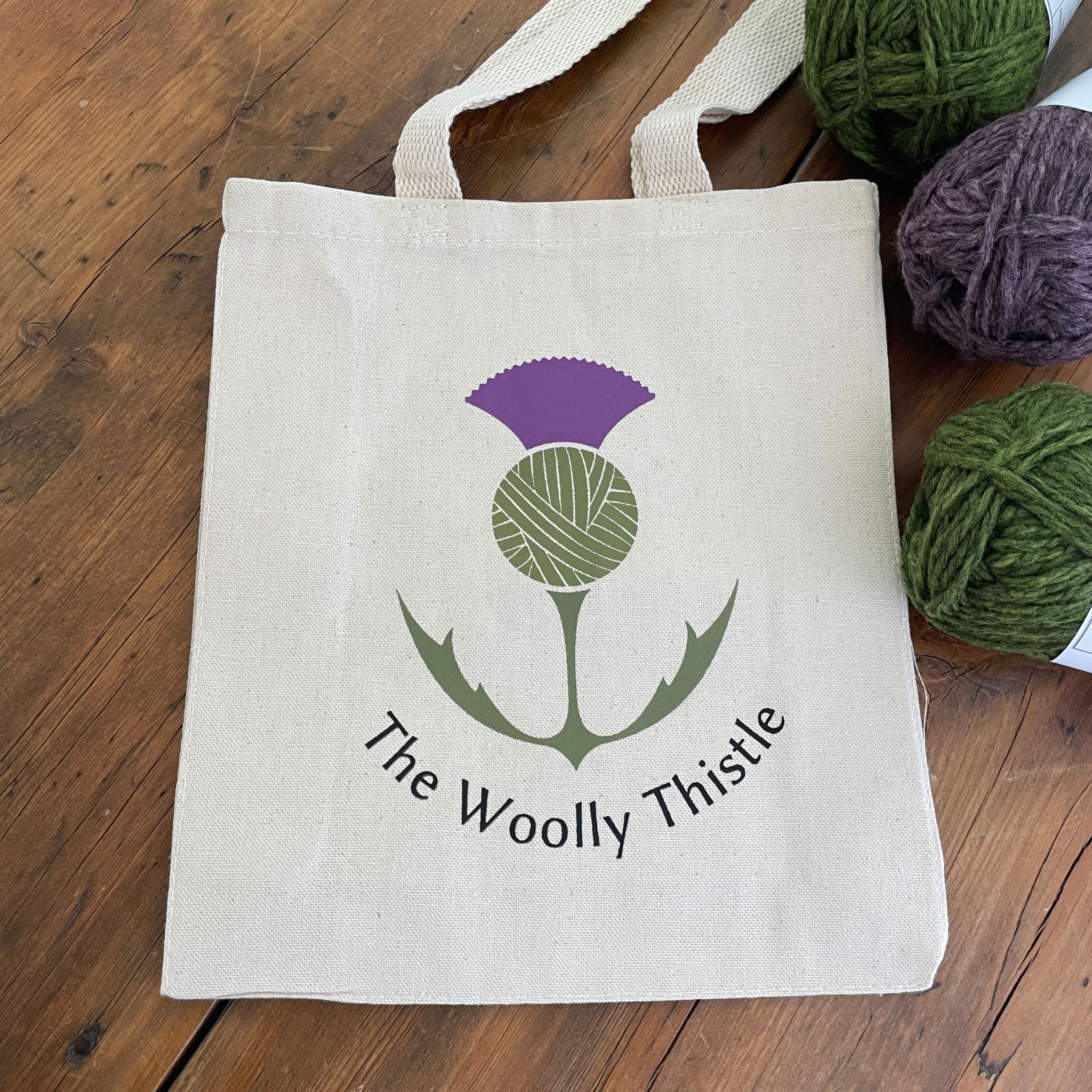 Canvas tote with The Woolly Thistle color logo.  Small size tote with green and purple yarn in background.