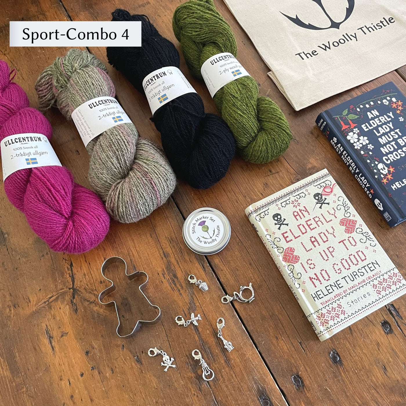 The Swedish Book Bundle components which include two books by Helene Tursten, TWT Tote bag, Stitch marker set, gingerbread cookie cutter, and Ullcentrum Sport Weight yarn. Components are shown and laid out on table. Yarn colors in Sport Combo 4 include a bright pink,  grey, black, and green.
