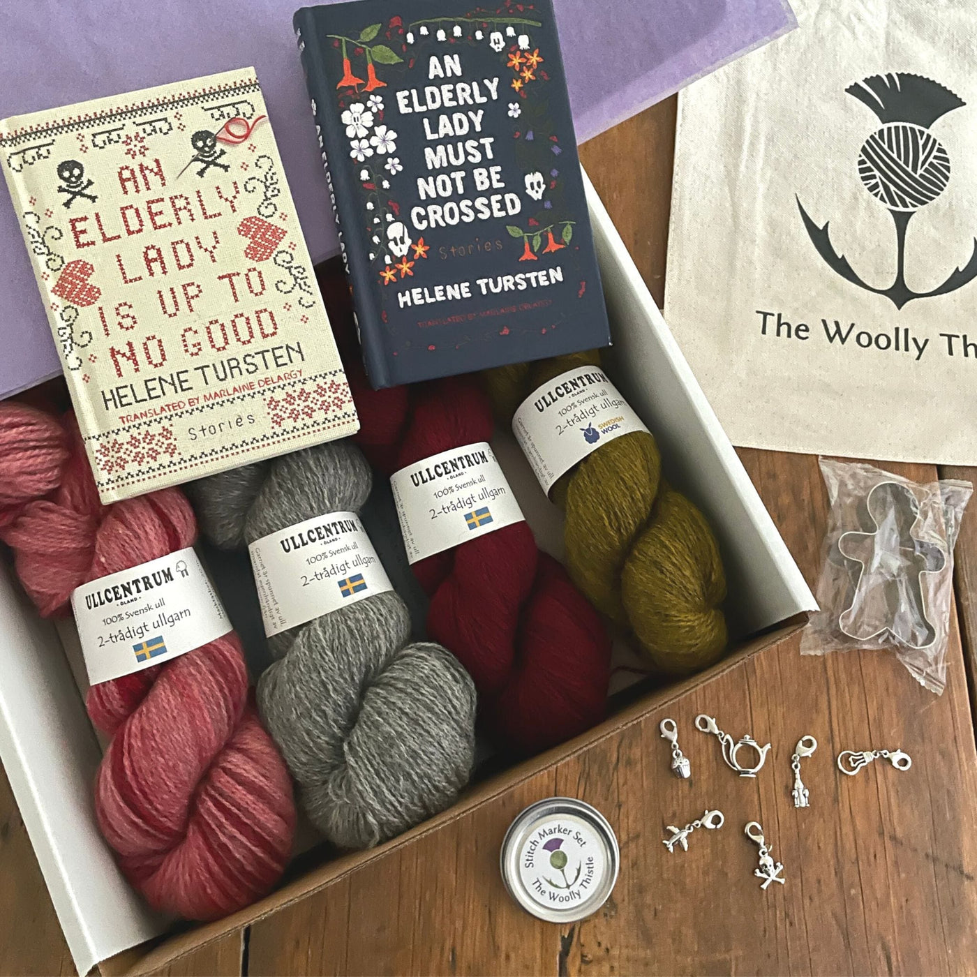 The Swedish Book Bundle components which include two books by Helene Tursten, TWT Tote bag, Stitch marker set, gingerbread cookie cutter, and Ullcentrum Sport Weight yarn. Components are shown partially in box and laid out on table. 