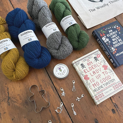 The Swedish Book Bundle components which include two books by Helene Tursten, TWT Tote bag, Stitch marker set, gingerbread cookie cutter, and Ullcentrum Worsted Weight yarn. Components are shown and laid out on table.