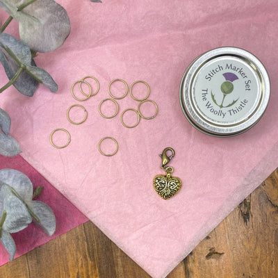 The Woolly Thistle tin shown with a golden heart stitch marker charm and stitch markers on pink background.