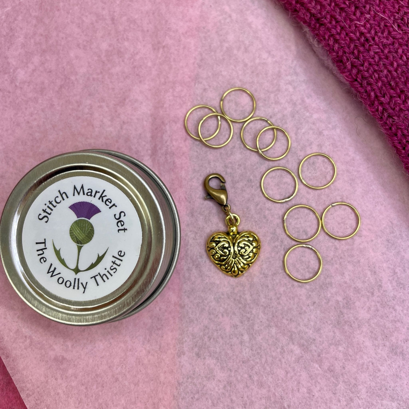 The Woolly Thistle tin shown with a golden heart stitch marker charm and stitch markers on pink background.