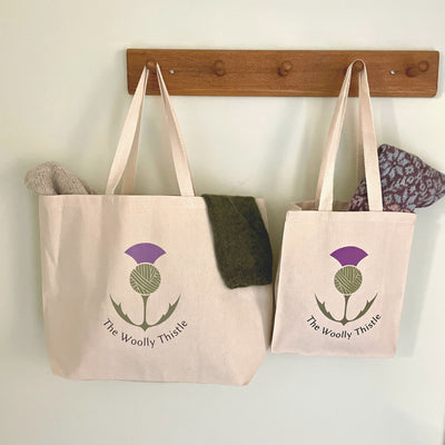 The Woolly Thistle color logo, canvas totes hanging on hooks. Large and small tote with knitted items peeking out.