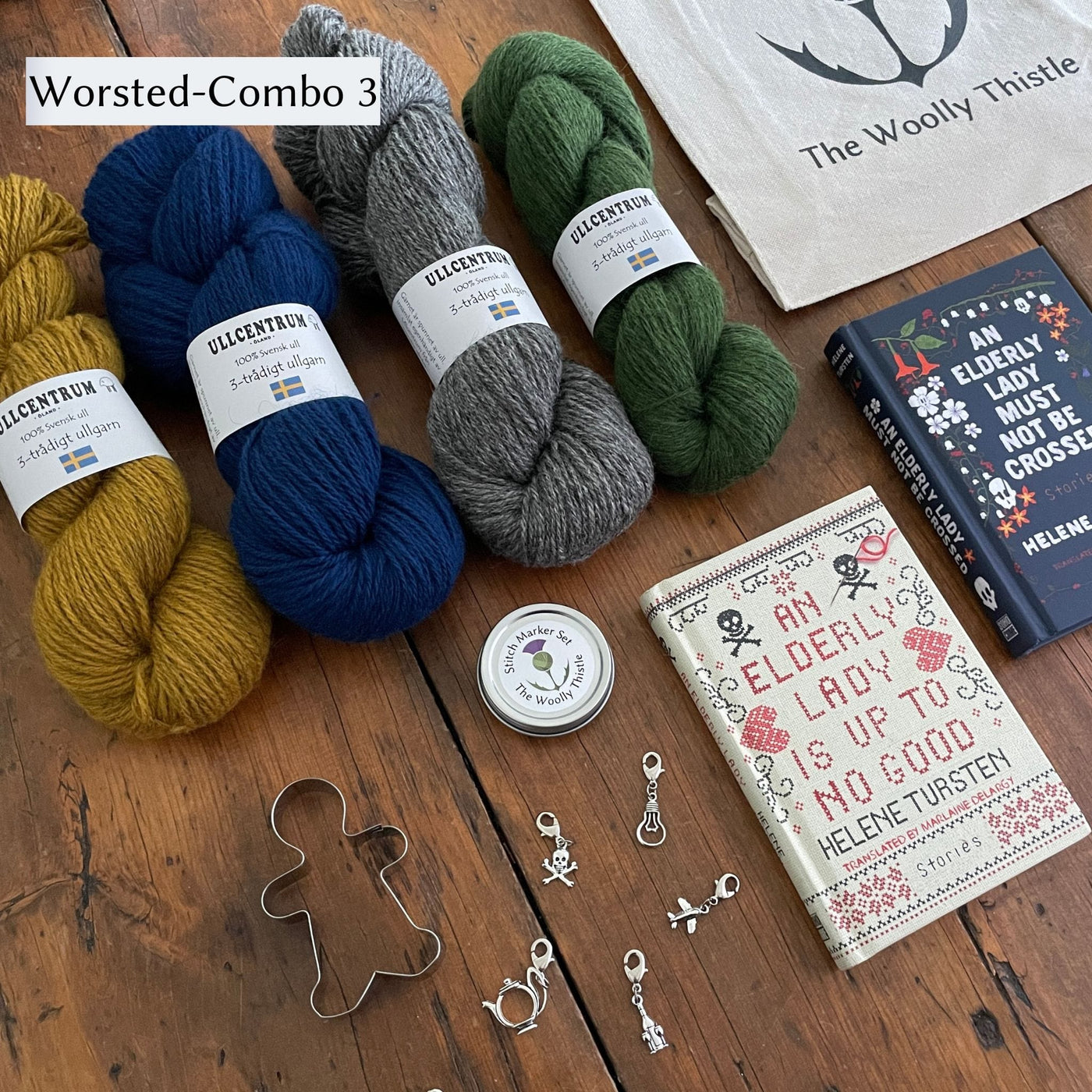 The Swedish Book Bundle components which include two books by Helene Tursten, TWT Tote bag, Stitch marker set, gingerbread cookie cutter, and Ullcentrum Worsted Weight yarn. Components are shown and laid out on table. Worsted Combo 3 includes Yarn in gold, blue, grey, and green. 