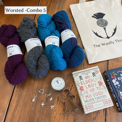 The Swedish Book Bundle components which include two books by Helene Tursten, TWT Tote bag, Stitch marker set, gingerbread cookie cutter, and Ullcentrum Worsted Weight yarn. Components are shown and laid out on table. Yarn colors included in Worsted Combo 5 include violet, grey, turquoise, and variegated blue. 