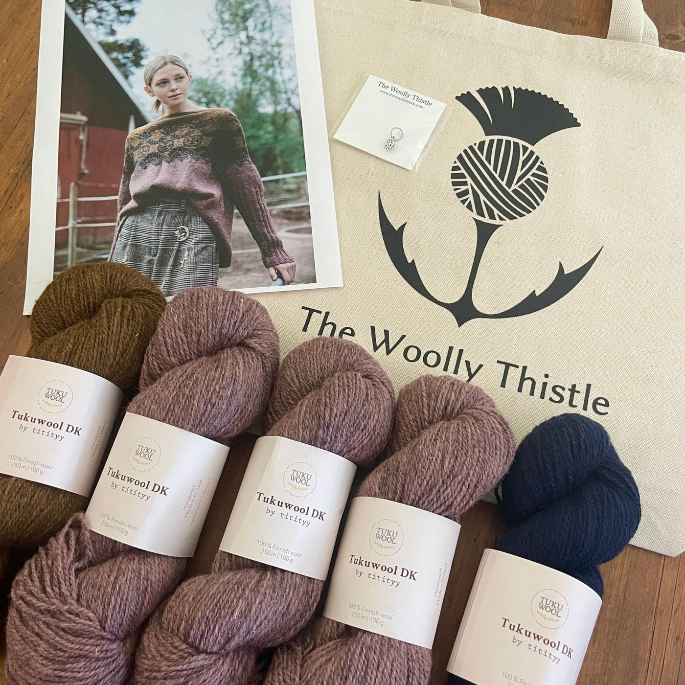 Components of the Pohjolan Neito Sweater yarn set: 5 skeins of Tukuwool DK in colors Ujo, Nila, and Tynni shown with photo of sweater and The Woolly Thistle tote bag and stitch marker. 
