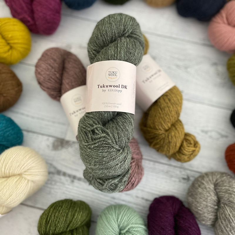 Tukuwool DK Yarn. Three skeins in heathered mauve, grey/green, and gold, framed by the end of many colorful skeins arranged in circle.