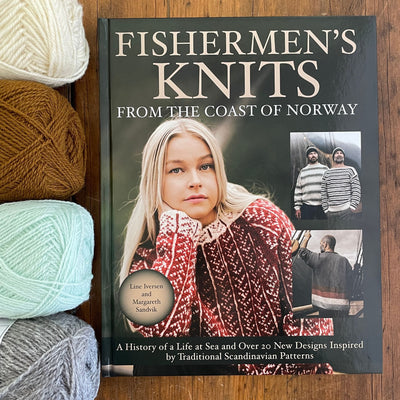 Fishermen's Knits from the Coast of Norway cover showing models wearing handknit sweaters. Book on wooden table next to yarn lined up on the left. 