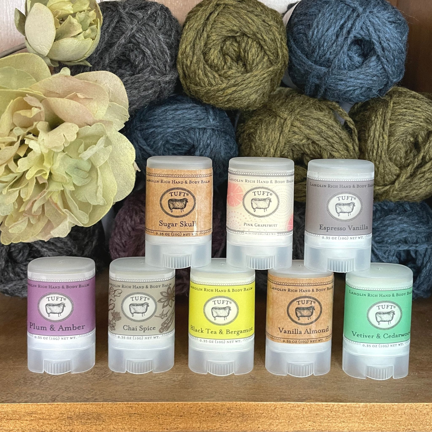 Eight containers of Tuft Woolens hand & body balm, in various scents, arranged on shelf in front of yarn. 