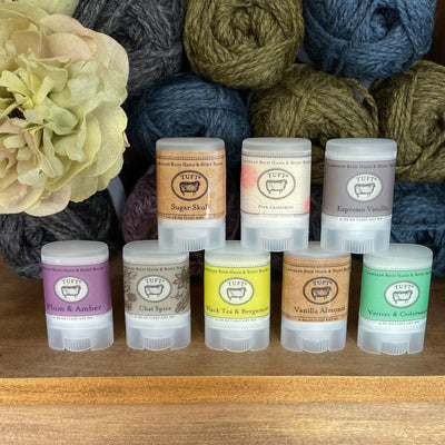 Eight containers of Tuft Woolens hand & body balm, in various scents, arranged on shelf in front of yarn. 