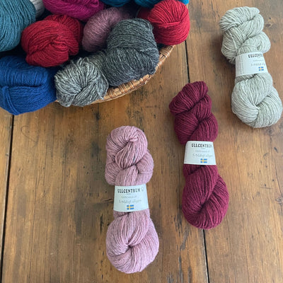 Three skeins of Ullcentrum 3ply, a worsted weight wool yarn, on a table, in pink, deep pink, and light grey on a table with a basket of other skeins nearby