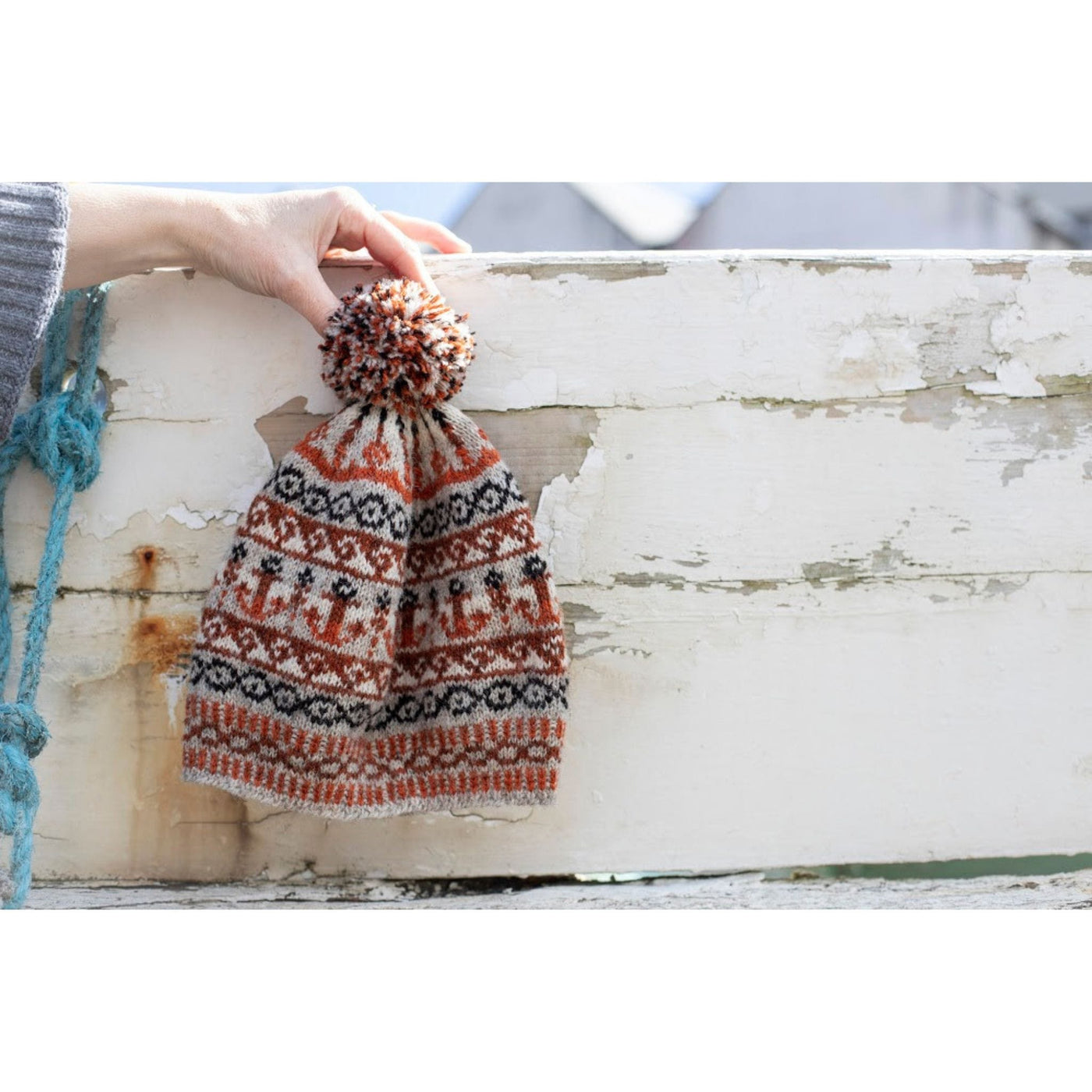 The Bonnie Isle Hat is being held agains a white background. This hat uses 6 cakes of Uradale 2ply Jumper Weight Yarn in Neutrals and two shades of orange.