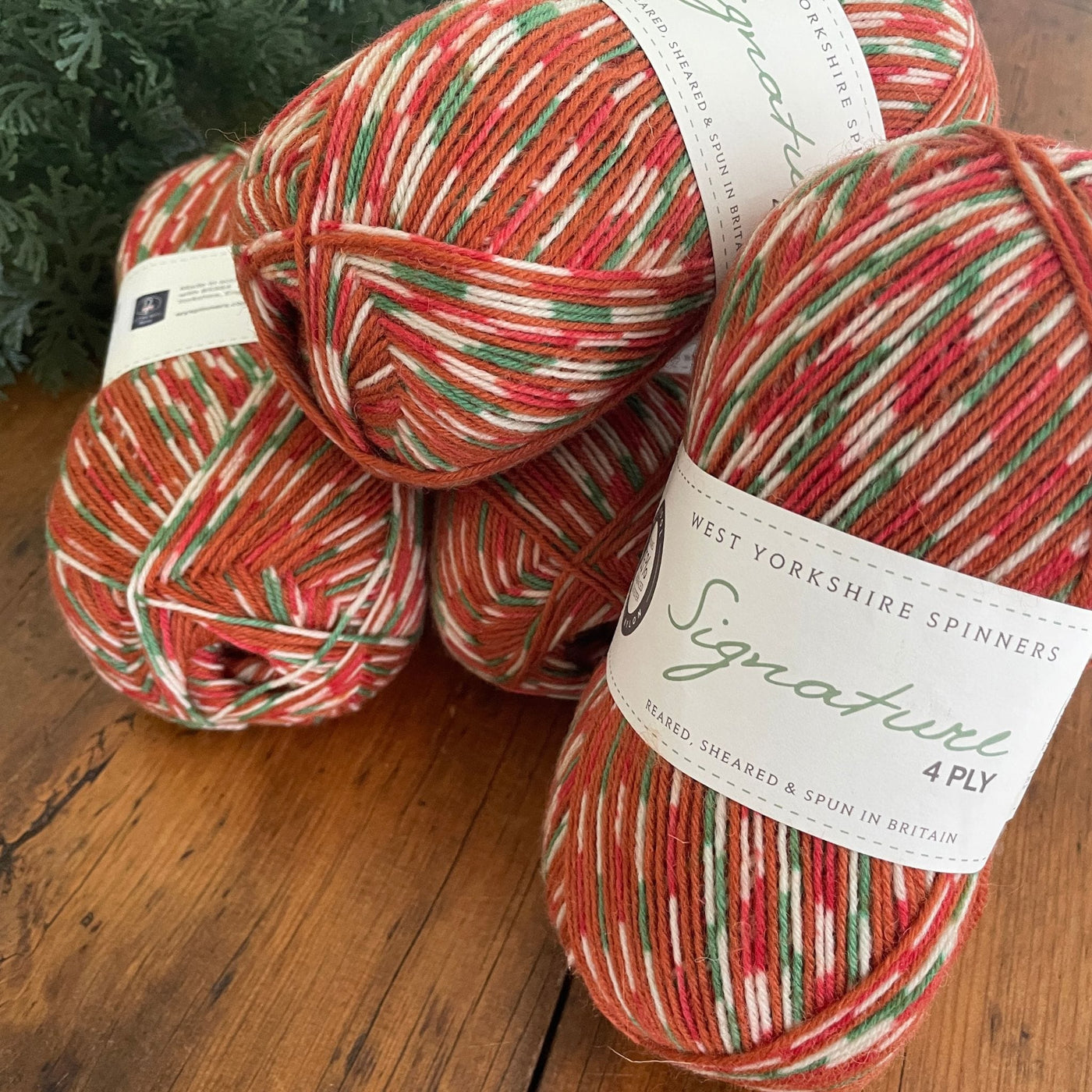 Four balls of West Yorkshire Spinners fingering weight yarn in Gingerbread, a warm brown, white, green, and red colorway