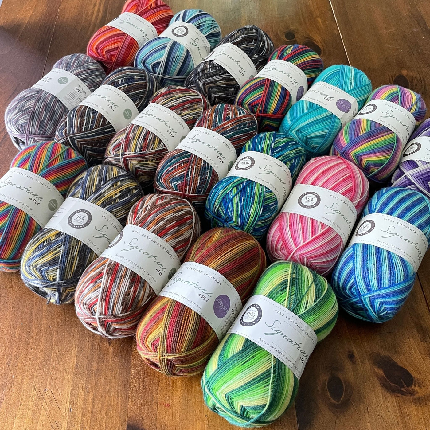 Three rows of West Yorkshire Spinners Signature 4ply yarn, fingering weight, in 100 gram balls, in striped or multicolored colorways, on a wooden table. 