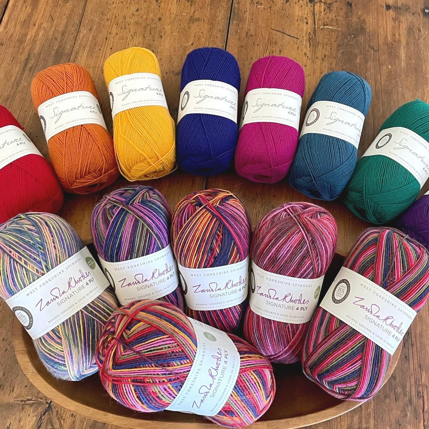 West Yorkshire Spinners Signature 4ply yarn, fingering weight, in 100 gram balls, arranged with seven solid colors on a table and six coordinating striped colors in a wooden bowl.