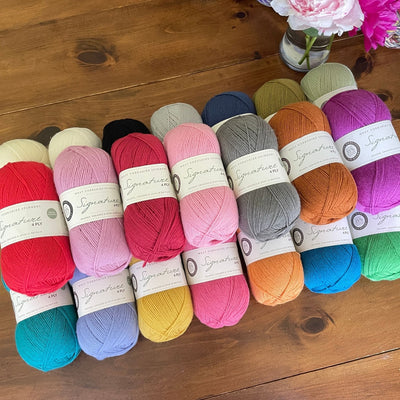 Stack of three rows of 100-gram balls of West Yorkshire Spinners Signature 4ply yarn in numerous solid colors on a wooden table near a glass vase with two pink flowers