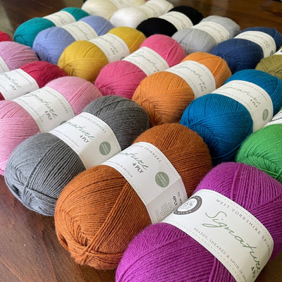 Numerous solid colors of 100-gram balls of West Yorkshire Spinners Signature 4ply  yarn laying in rows on a wooden table