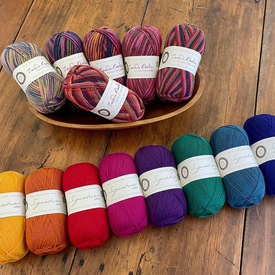 Eight 100-gram balls of solid color West Yorkshire Spinners Signature 4ply yarn (1002 Fuschia, 1000 Rouge, 1004 Amber, 1003 Amethyst, 1005 Cobalt, 1006 Spruce, 1001 Sunflower, and 1007 Pacific) on a wooden table near a wooden bowl of coordinating self-striping yarns. 